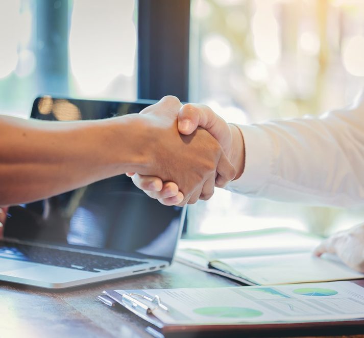 Two people shaking hands, with a laptop and paper work on a table