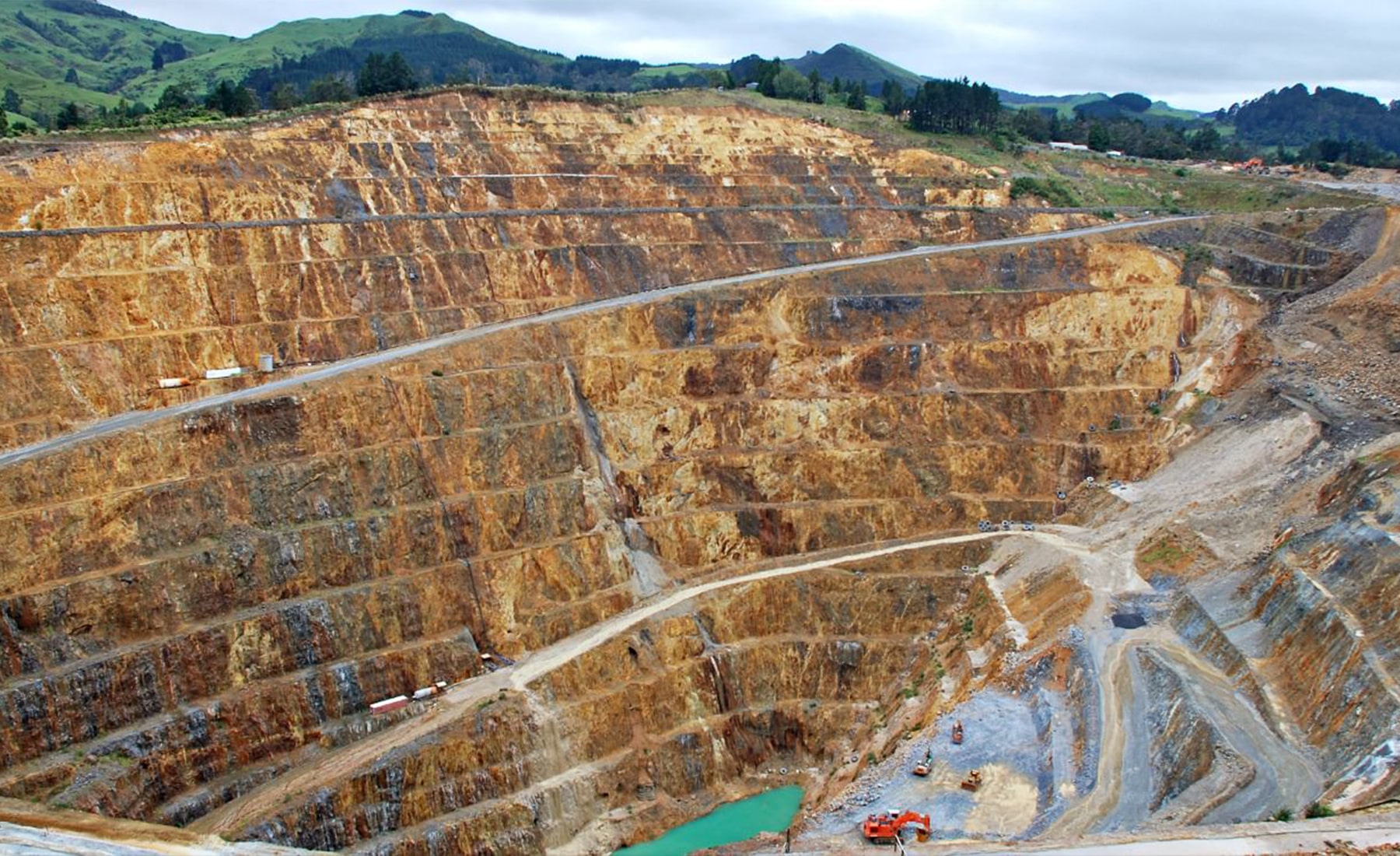 Deep mining quarry, with construction machinery throughout. At the bottom of the quarry lies a small lake of water.