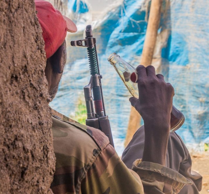 Solider sat down by tree, drinking a bottle of soft drink, whilst holding a gun