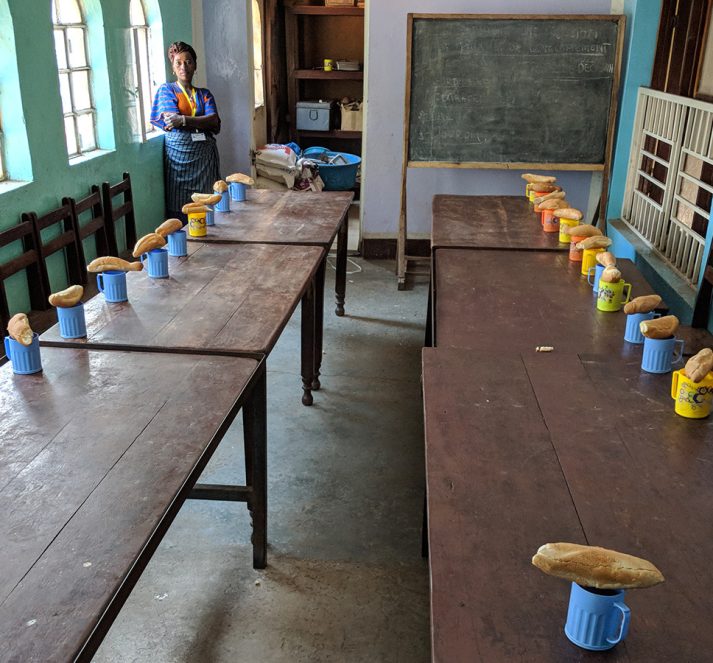 A school classroom, with mugs and bread on the desks. Chalkboard and teacher in the background