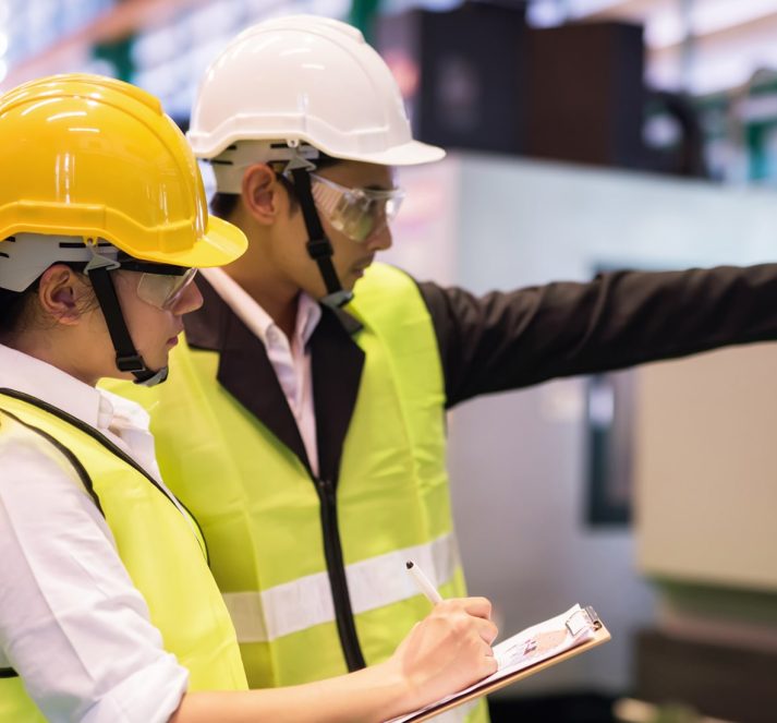 Male and female in a factory, wearing safety hats and hi vis jackets. Male pointing into the distance.