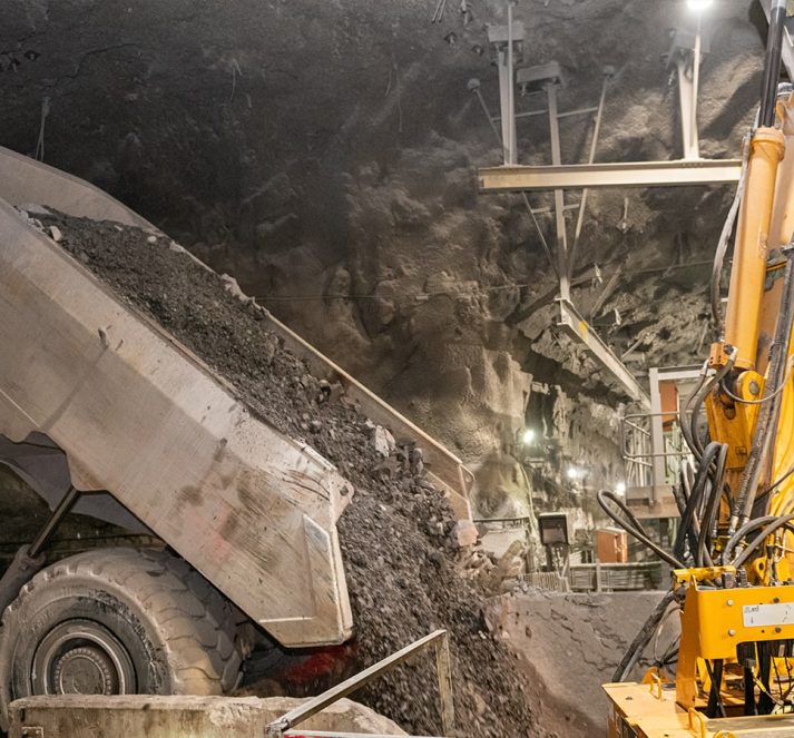 Large truck dropping mined materials next to yellow machinery inside a mine