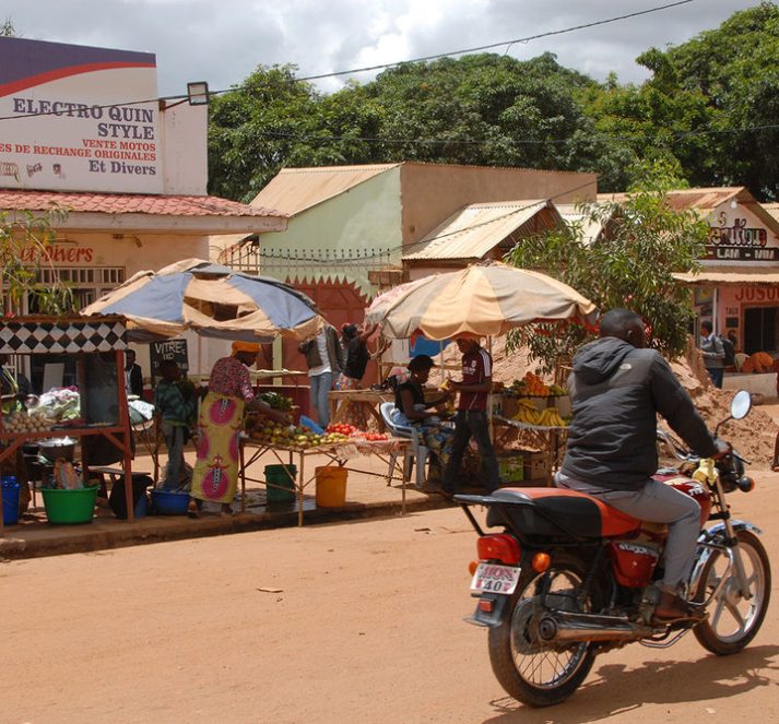 Man on a motorbike in the foreground of a town, with stalls selling fruit and vegetables